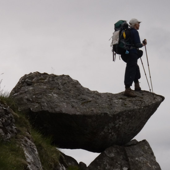 Zoom: Lady with rucksack and camerabag watching evening clouds from a balancing rock on Meall nan Eun